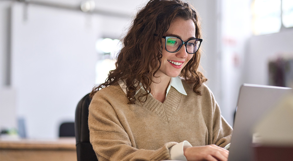 Young woman wearing glasses sitting at a desk looking at a computer.