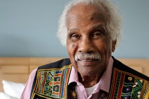 Headshot of Ashley Bryan; an older African American wears a kenti cloth vest and looks into the camera.